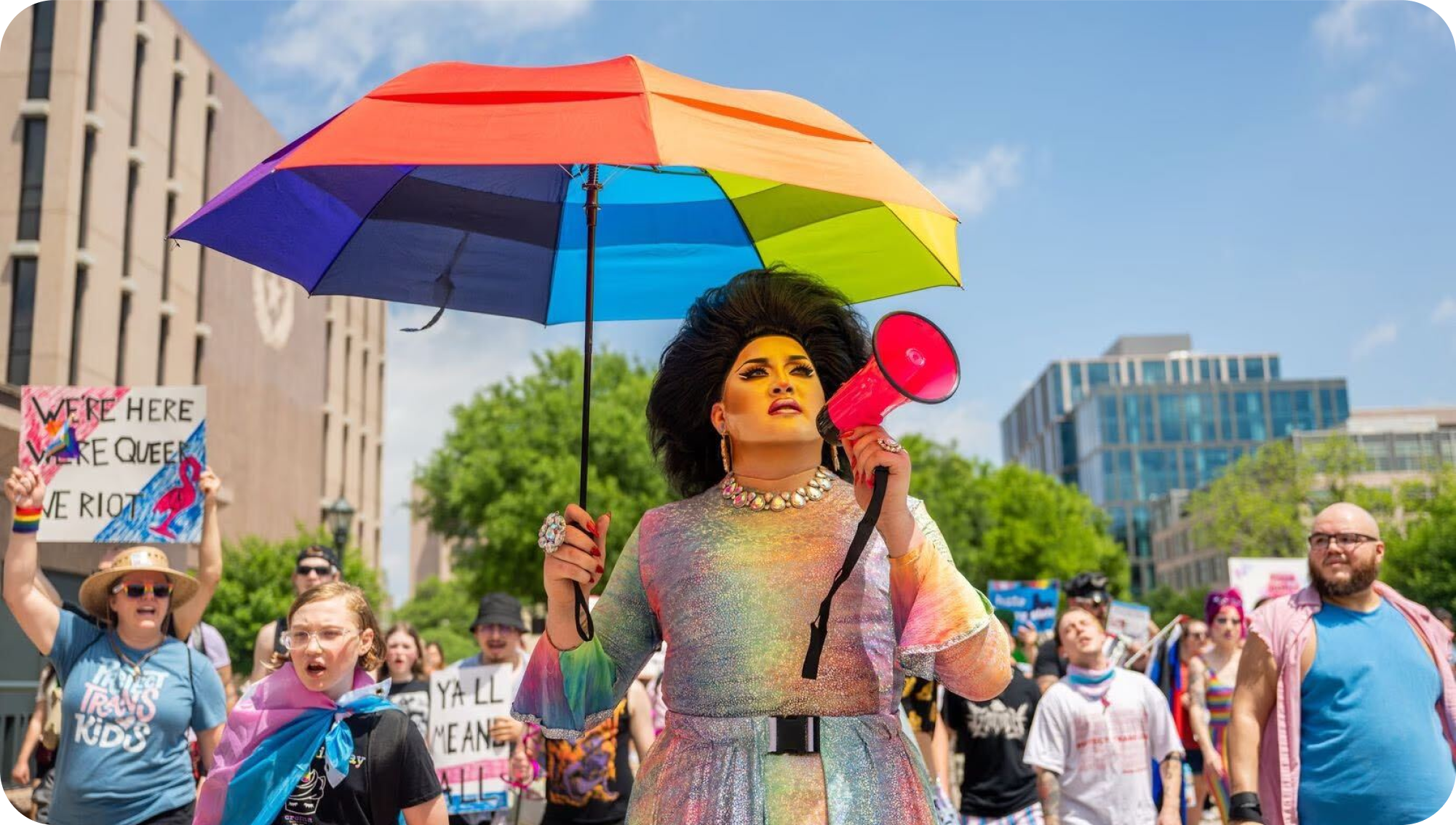 A drag queen holding a megaphone in one hand and a rainbow umbrella in the other in front of a group of people holding signs and marching together.