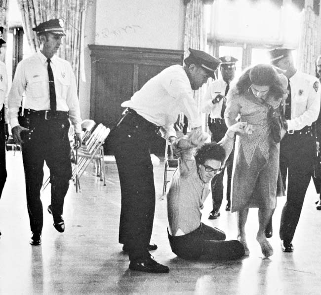 A black and white photo from 1970 of two people being dragged by police officers.