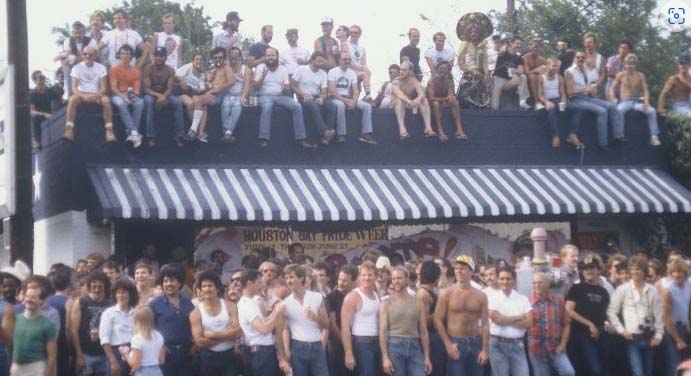 A color photo from 1971 of a large crowd of spectators with some standing in front of a building and some sitting on the roof of the building.