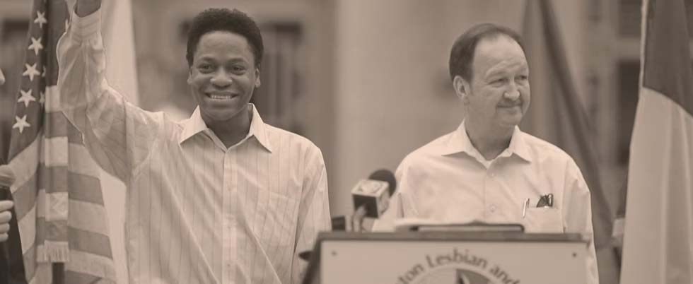 A sepia photo from 2003 of two men standing behind a podium. One of the men is waving and smiling.