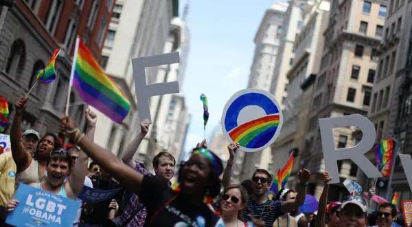 A color photo from 2012 of a crowd of people marching in the street cheering and holding pride flags and posters.