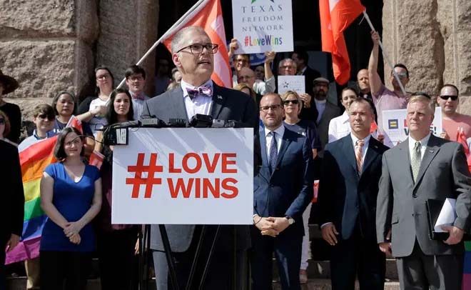 A color photo from 2015 of a man standing at a podium with a sign that reads "#Love Wins." Behind him a small crowd of people holding pride flags and posters look on.