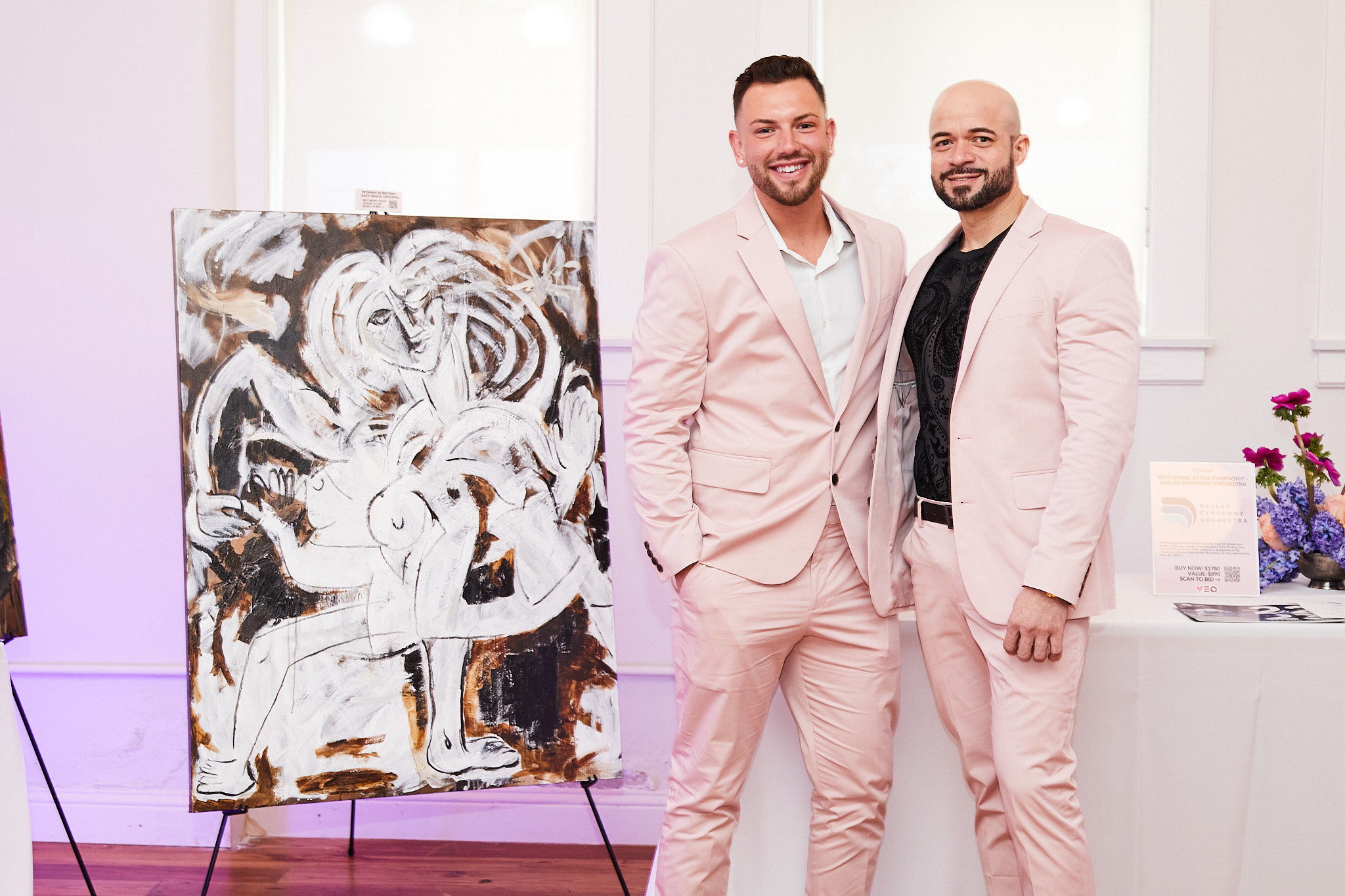 2 men wearing light pink suits standing next to a painting and smiling at the camera.