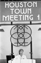 A black and white photo of a woman sitting below a sign that reads "Houston Town Meeting 1"